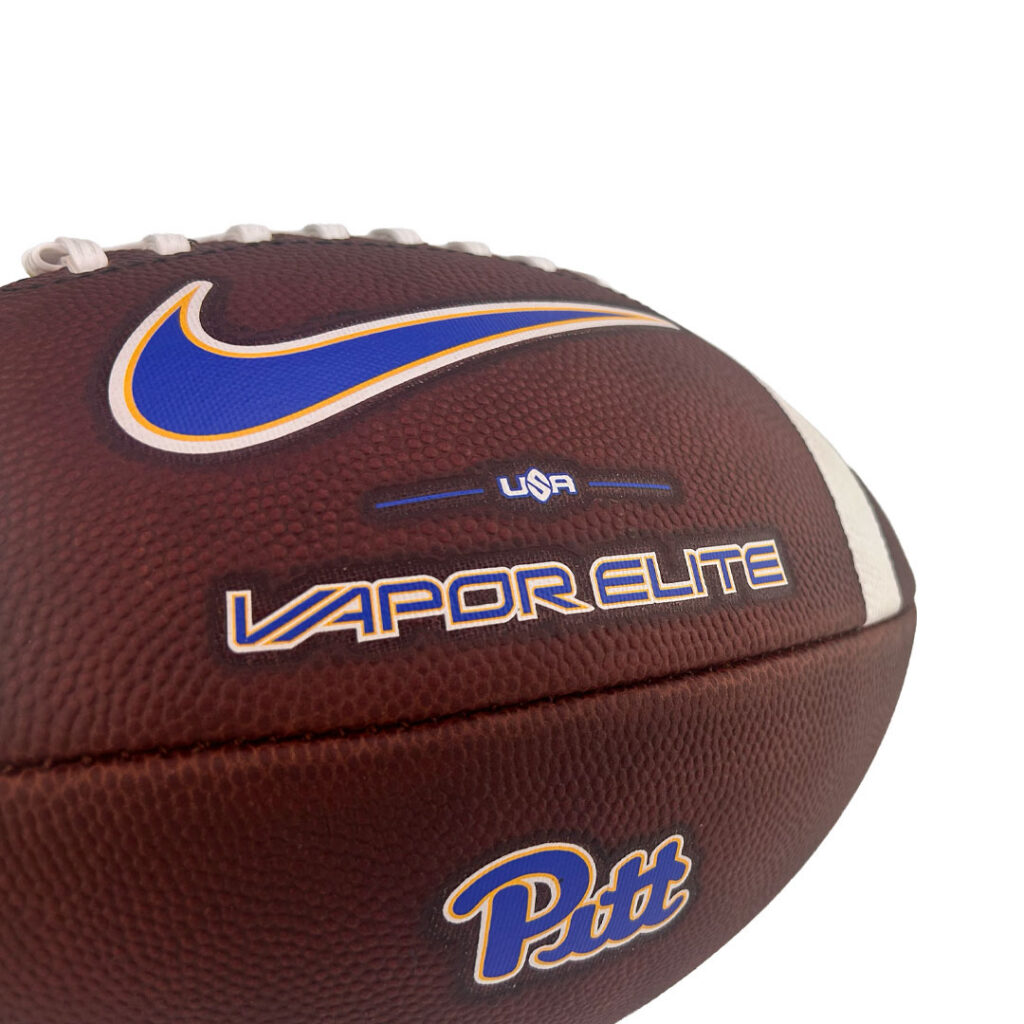 Close-up of a brown football with white laces, displaying a blue and orange Nike swoosh and the words 'USA' and 'VAPOR ELITE' along with a Pitt logo on the lower panel.
