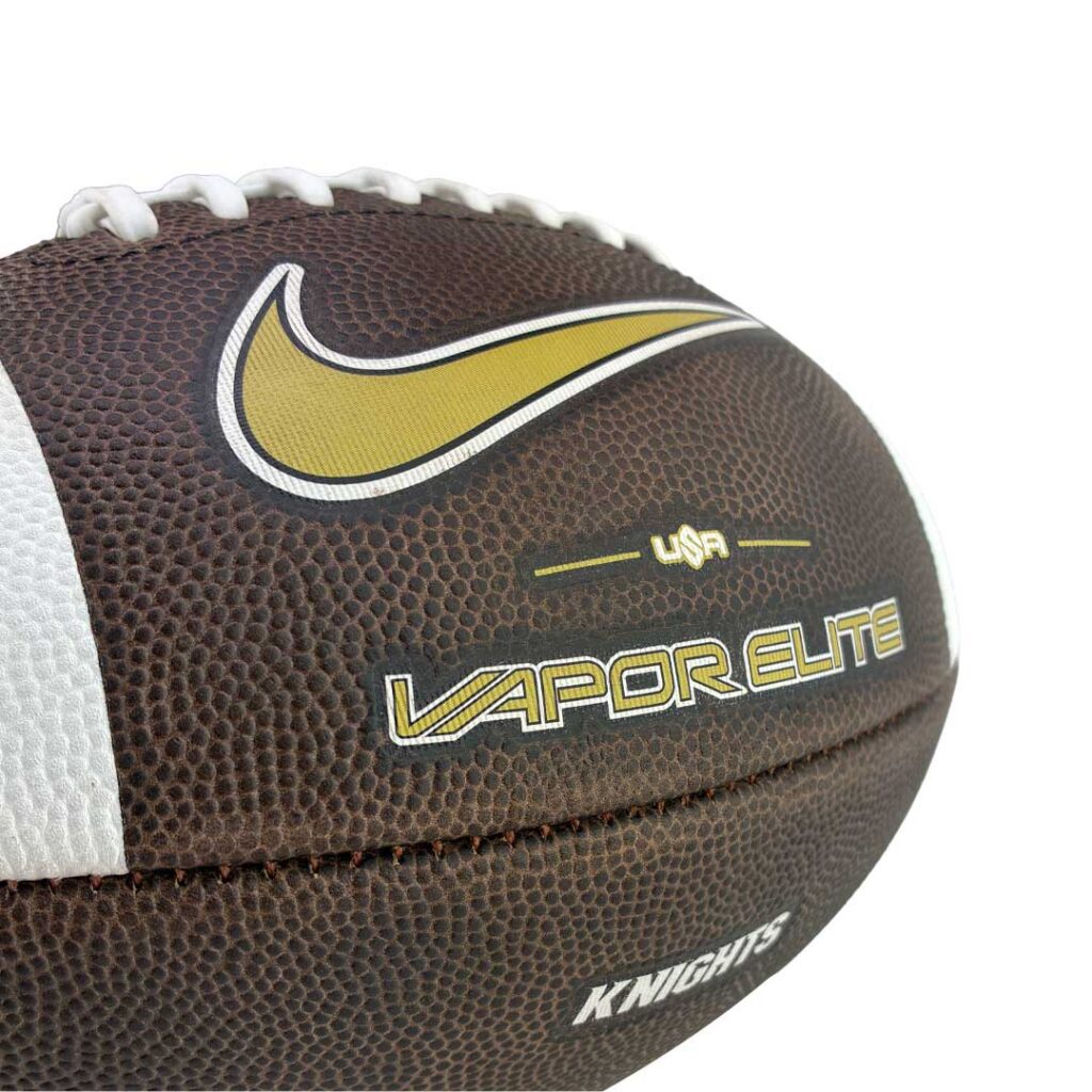 Close-up of a brown football with white laces, featuring a gold Nike swoosh and the 'Vapor Elite' branding.
