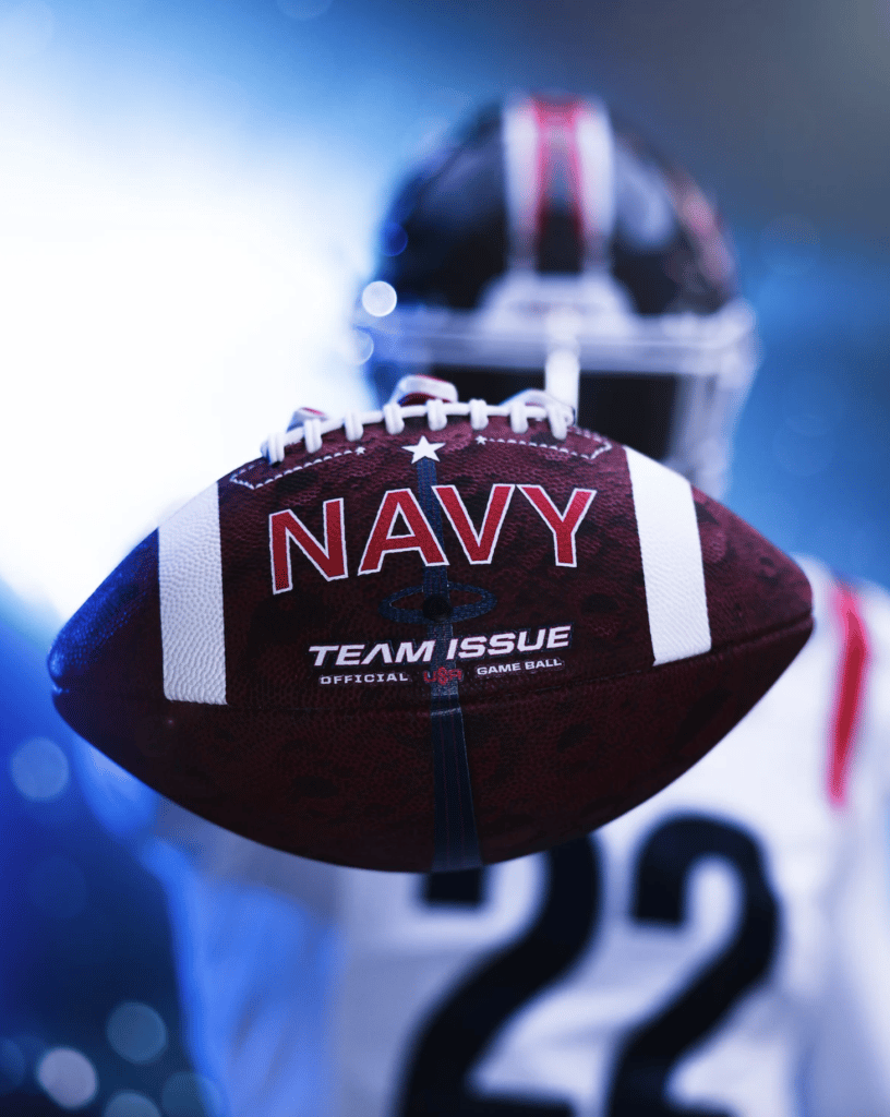 Image of Navy football player in uniform blurred in the background, holding Navy football close to the camera in focus