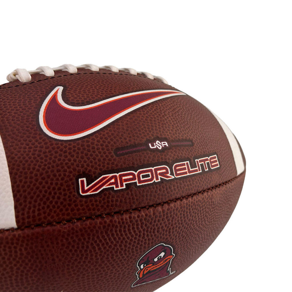 Close-up of a brown leather football with white laces, a red Nike swoosh, 'USA' and 'VAPORELITE' branding, plus a small mascot patch.