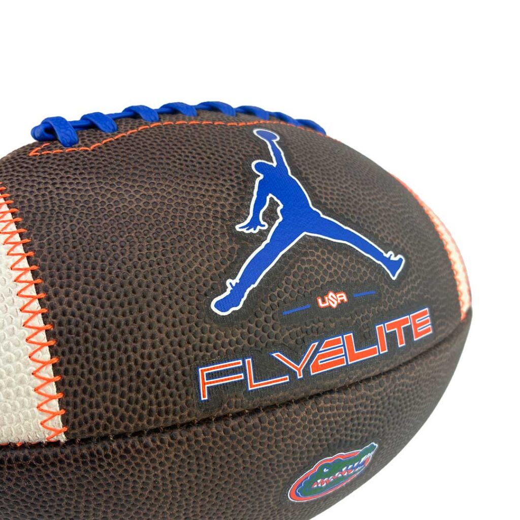 Close-up of a brown textured football with blue laces, featuring a white-blue Jumpman silhouette and the word FLYELITE with a Florida Gators patch nearby