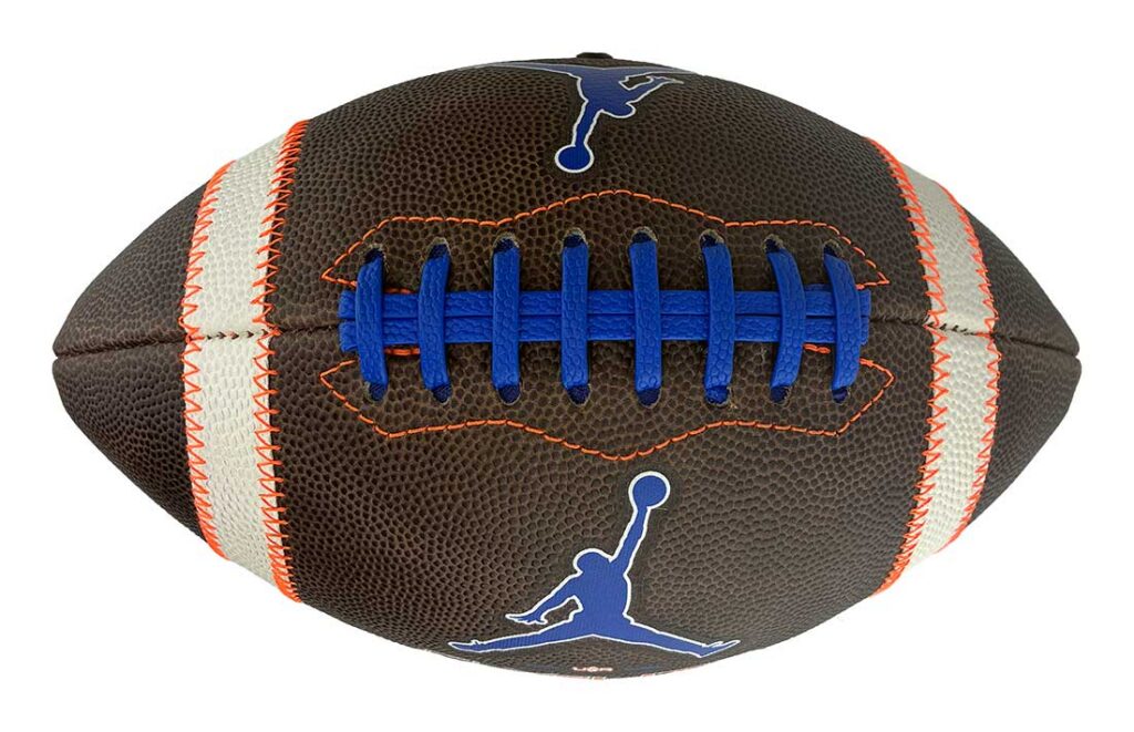 Football with blue laces and Jumpman logos on a brown leather surface, white stripes with orange stitching along the seams, center-focused view.