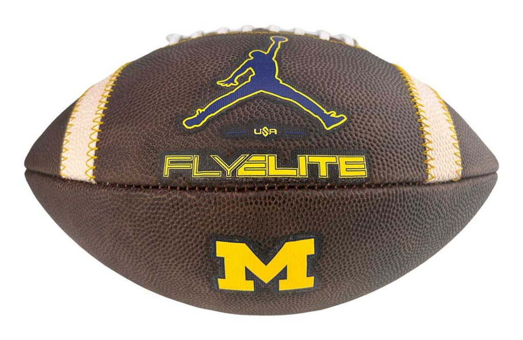 Brown football with yellow Michigan 'M' logo, yellow seams, and 'Flyelite' branding.