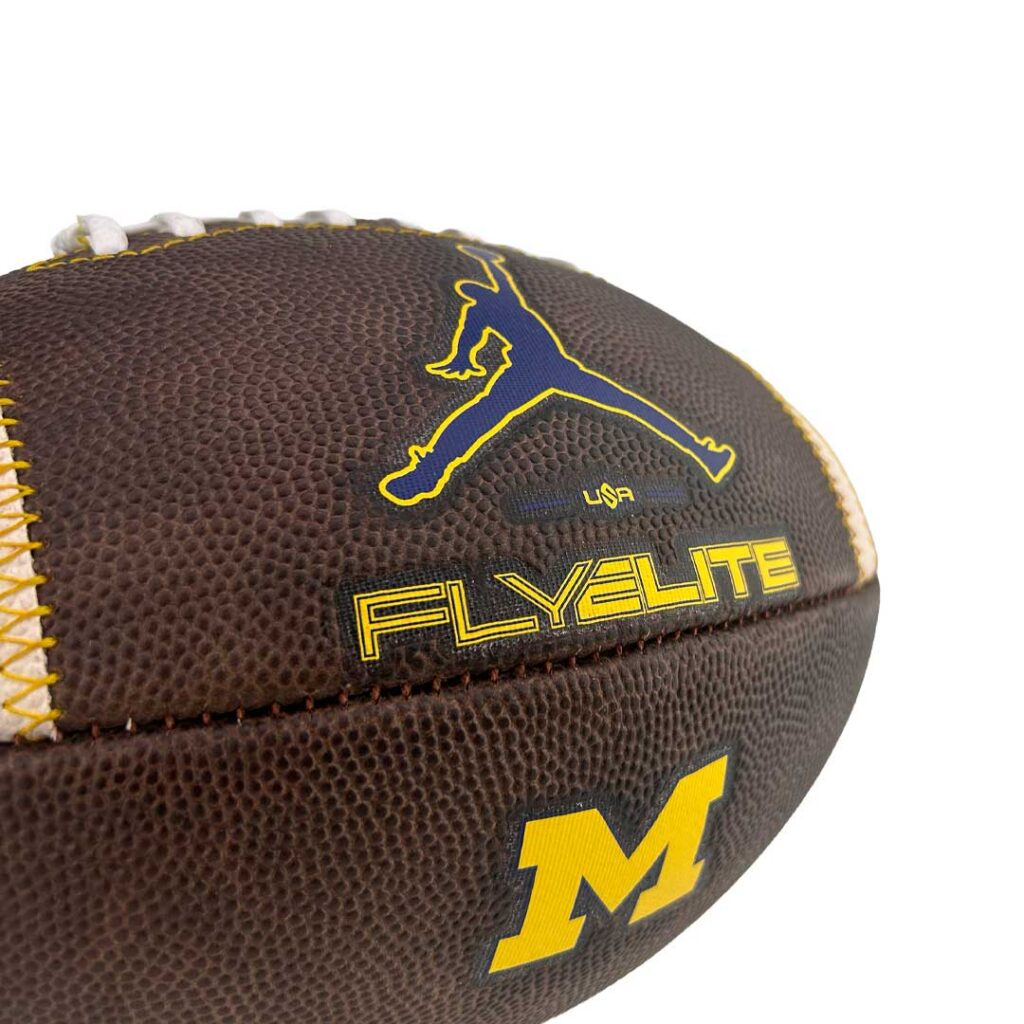 Brown football close-up featuring yellow stitching, a blue Jumpman logo, 'FLYELITE' in yellow, and a yellow Michigan 'M' logo nearby.