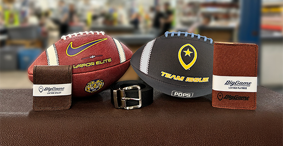 Three branded footballs and two leather belts arranged on a brown display table in a sports store, with blurred shoppers in the background.