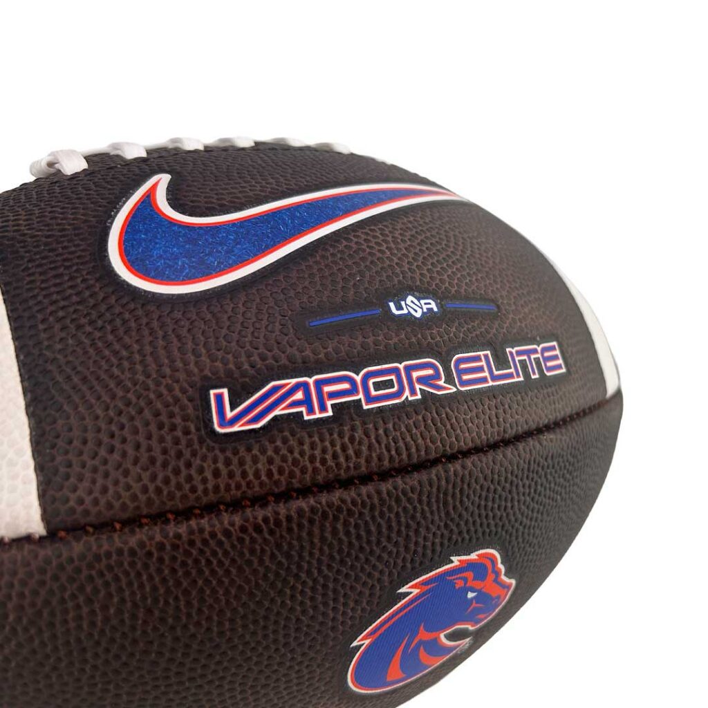Close-up of a brown football with blue and red Nike logo and 'VAPOR ELITE' text, showing a USA branding and team emblem on the side